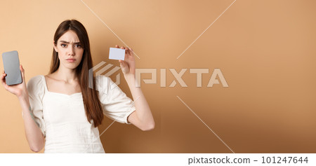 Sad and disappointed girl showing empty smartphone screen and plastic credit card, complaining and frowning, standing on beige background Sad and disappointed girl showing empty smartphone screen and plastic credit card, complaining and frowning, standing on beige background 101247644