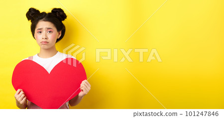 Heartbroken asian woman showing big red heart cutout and looking sad, feeling lonely on lovers day, showing valentine card at camera, yellow background 101247846