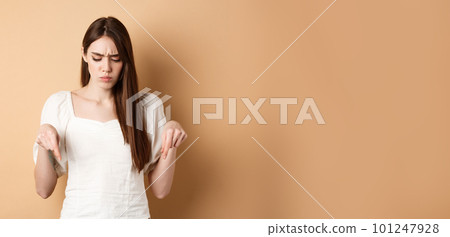 Upset young woman frowning and looking down, pointing at bad thing, standing confused and displeased on beige background Upset young woman frowning and looking down, pointing at bad thing, standing confused and displeased on beige background 101247928