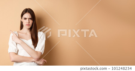 Angry frowning girl pointing left at empty space, looking disappointed and upset, scolding you or accuse, standing on beige background 101248234