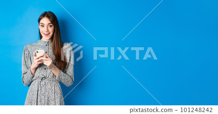 Cute smiling girl with long natural hair, wearing dress, using smartphone and looking happy, standing against blue background Cute smiling girl with long natural hair, wearing dress, using smartphone and looking happy, standing against blue background 101248242