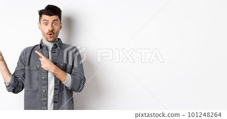Excited man pointing left at logo and look with disbelief, saying wow at camera, checking out cool promo discount, standing on white background 101248264