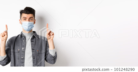 Pandemic lifestyle, healthcare and medicine concept. Stylish modern man in medical mask showing advertisement, pointing fingers up at logo, standing on white background 101248290