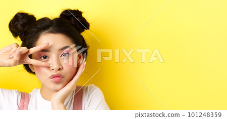 Close-up of cute asian woman with bright glamour makeup, long fingernails, showing v-sign and pouting silly at camera, standing kawaii on yellow background 101248359