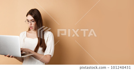Worried girl look at laptop screen, typing on computer with focused face. Woman freelancer working in internet, standing on beige background 101248431