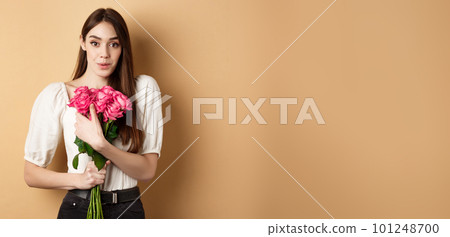 Valentines day. Surprised tender girl thanking for flowers and smiling, holding red roses and smiling grateful, standing on beige background 101248700