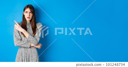 Confused girl in dress pointing and looking at upper left corner with puzzled frowning face, standing hesitant on blue background Confused girl in dress pointing and looking at upper left corner with puzzled frowning face, standing hesitant on blue background 101248734