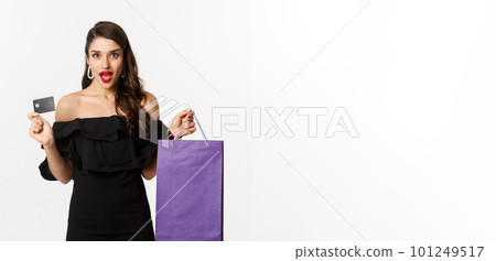 Stylish young woman in black dress going shopping, holding bag and credit card, smiling pleased, standing over white background 101249517