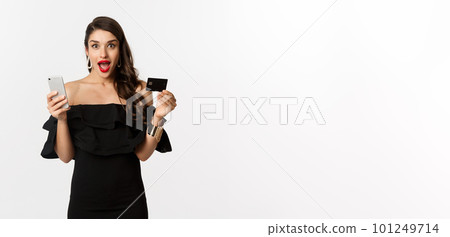 Online shopping concept. Fashionable woman in black dress, holding credit card with smartphone, looking excited, standing over white background 101249714