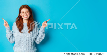 Winter holidays and people concept. Cute teen girl with red hair, smiling and pointing fingers sideways, showing advertisements, standing over blue background 101249825