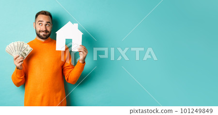 Real estate concept and mortgage concept. Handsome young man buying house, holding home maket and money, thinking and smiling, standing over light blue background 101249849