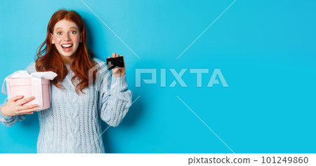 Winter holidays promo offer concept. Cheerful redhead woman holding Christmas gift and credit card, staring at camera amazed, standing over blue background 101249860
