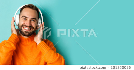 Close-up of handsome modern man listening music in headphones, standing in orange sweater over turquoise background Close-up of handsome modern man listening music in headphones, standing in orange sweater over turquoise background 101250056