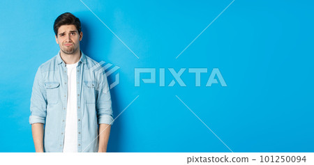 Man looking with doubt at camera, frowning and sulking, feeling uneasy about something, standing against blue background 101250094