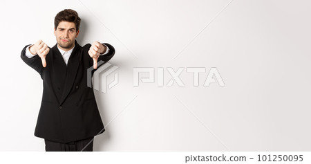 Portrait of skeptical and disappointed man in black suit, frowning upset, showing thumbs-down, dislike something bad, standing over white background 101250095