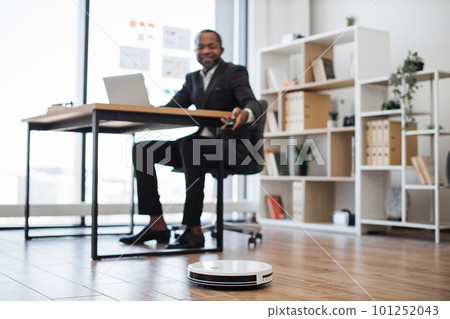 Robotic vacuum cleaning office while man working on laptop Robotic vacuum cleaning office while man working on laptop 101252043