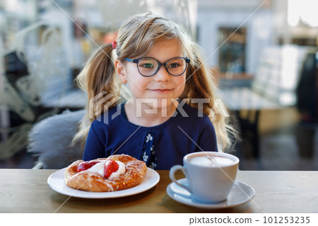 Adorable smiling girl with glasses have a breakfast in a cafe. Preschool child with glasses drinking chocolate and eating bakery pastry croissant or cake. Happy children, healthy food and meal. 101253235
