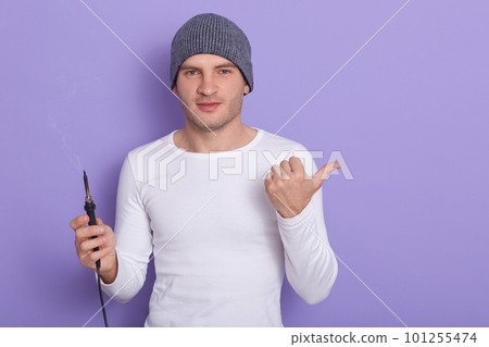 Young technician being ready to solder wire, attractive male wears white casual shirt and gray cap holds soldering iron in one hand and point aside with another thumb, isolated over purple background. Young technician being ready to solder wire, attractive male wears white casual shirt and gray cap holds soldering iron in one hand and point aside with another thumb, isolated over purple background. 101255474