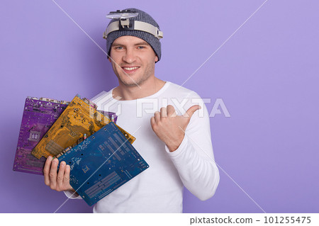 Happy smiling radiotrician wears casual outfit with magnifier on her forehead, holding three multicolored motherboards or flares in hand, showing something aside with his thumb. Electronic engineering Happy smiling radiotrician wears casual outfit with magnifier on her forehead, holding three multicolored motherboards or flares in hand, showing something aside with his thumb. Electronic engineering 101255475