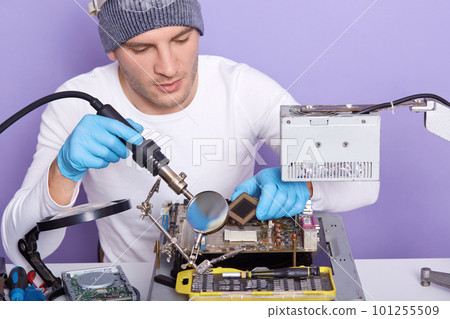 Horizontal shot of hardworking guy using soldering iron at work, male wearing white shirt, gray cap and blue rubber gloves, having concentrated look, radioman posing isolated over lilac studio wall. 101255509