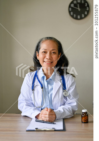 An Asian-aged female doctor in white gown sits at her desk in the hospital office. 101255879