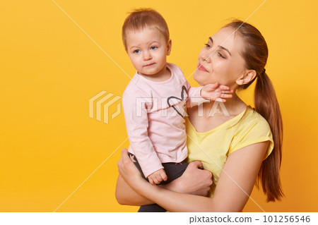 Studio shot of loving mother holding her baby girl, toddler looking aside, mom admires her charming doughter isolated over yellow studio wall. Motherhood, happyness, children concept. Copy space. 101256546
