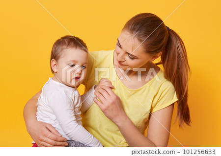 A beautiful woman on maternity leave and her cute little daughter hug each other on bright yellow background. A sweet child looks with interest aside. Family relationship between mother and daughter. A beautiful woman on maternity leave and her cute little daughter hug each other on bright yellow background. A sweet child looks with interest aside. Family relationship between mother and daughter. 101256633