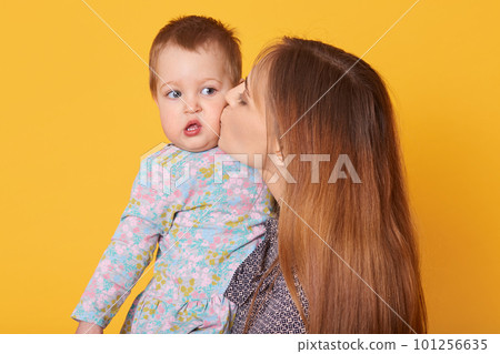 Indoor shot of beautiful tender woman holding her daughter, kissing her in cheek. Tiny cute little girl looks aside, opens her mouth with interest, wearing light blue dress. Family and care concept. Indoor shot of beautiful tender woman holding her daughter, kissing her in cheek. Tiny cute little girl looks aside, opens her mouth with interest, wearing light blue dress. Family and care concept. 101256635