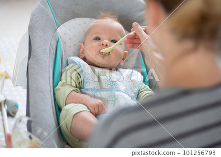 Mother spoon feeding her baby boy child in baby chair with fruit puree. Baby solid food introduction concept 101256793