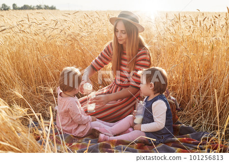 Caring helpful mother sitting on blanket at wheat field with her lottle daughters, filling glasses with milk from bottle, wearing dress and hat, childen behaving in proper way. Motherhood concept. 101256813