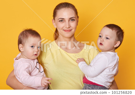 Image of mother with kids, two daughters in casual clothes, beautiful young woman with little twins standing in photo studio isolated over yellow background. Girls being interested to pose with mommy. 101256820