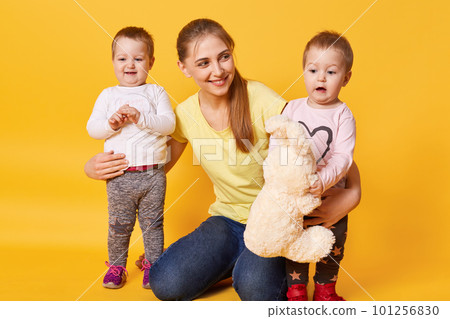 Imagine of young beautiful mother with little twin girls posing isolated over yellow background, children wants to play with their mommy, girls like to spend time with mum. Love and family concept. Imagine of young beautiful mother with little twin girls posing isolated over yellow background, children wants to play with their mommy, girls like to spend time with mum. Love and family concept. 101256830