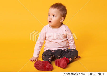 Studio shot of adorable little baby girl sitting on floor, isolated on yellow background, lovely baby portrait wearing casual clothes and red shoes, looks aside, looking for her mother. Copy space. 101257011