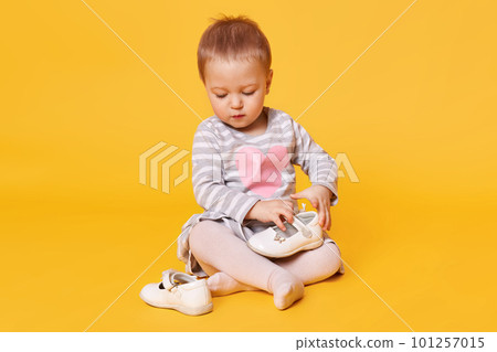 Fair haired pretty little child wearing stripped dress with heart poses isolated over bright yellow background. Sweet magnetic kid sits on floor in studio holding and observing her white shoes. 101257015