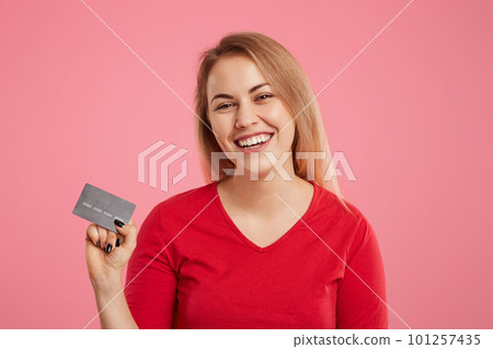 Isolated shot of cheerful pretty female holds plastic card in hands, wears red sweater, going to make online purchases, glad to recieve money bonus on her account, poses against pink background Isolated shot of cheerful pretty female holds plastic card in hands, wears red sweater, going to make online purchases, glad to recieve money bonus on her account, poses against pink background 101257435