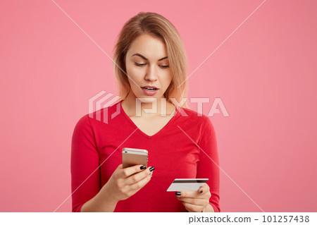Indoor shot of puzzled female doesn`t recieve salary, checks bank account via online application, uses modern cell phone and plastic card, types password, isolated on pink background. Online shopping Indoor shot of puzzled female doesn`t recieve salary, checks bank account via online application, uses modern cell phone and plastic card, types password, isolated on pink background. Online shopping 101257438