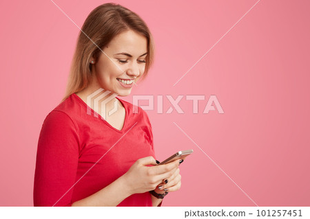 Smiling pleasant looking woman in red outfit holds smart phone, searches social networks, has short hairstyle, connected to wireless internet, isolated over rosy background with empty space for text Smiling pleasant looking woman in red outfit holds smart phone, searches social networks, has short hairstyle, connected to wireless internet, isolated over rosy background with empty space for text 101257451