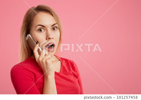 Shocked puzzled young female has phone conversation via telephone, surprised to hear unexpected news, isolated over pink background with copy space for your advertisment or promotional text. 101257458
