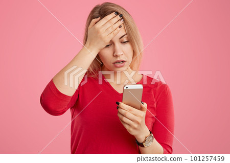 Stressful young lady keeps palm on forehead, reads shocking news in internet, focused in mobile phone, wears red casual jumper, isolated over pink background. Troublesome woman poses indoor. Stressful young lady keeps palm on forehead, reads shocking news in internet, focused in mobile phone, wears red casual jumper, isolated over pink background. Troublesome woman poses indoor. 101257459