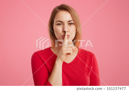 Portrait of secret beautiful young woman keeps fore finger on lips, shows silence sign, tries to keep personal information in secret, isolated over pink background. Hush or stop talking, please 101257477