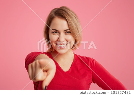 Smiling pretty young female has broad shining smile indicates at you with fore finger, makes choice, glad to be photographed, isolated over pink background. Facial expressions and gestures 101257481