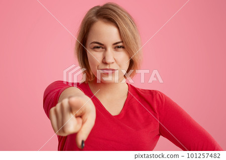 Suspicious young woman with short hair, points directly a camera, suspects someone, dressed in red clothes, poses over pink background, has pleasant apperance, isolated over pink background. Suspicious young woman with short hair, points directly a camera, suspects someone, dressed in red clothes, poses over pink background, has pleasant apperance, isolated over pink background. 101257482