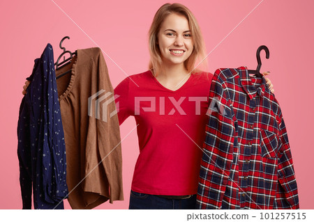 Isolated shot of happy blonde woman with toothy smile, chooses new outfit in shop, holds hangers with shirts, being in good mood, likes shopping, dressed in red jumper, poses over pink wall. Isolated shot of happy blonde woman with toothy smile, chooses new outfit in shop, holds hangers with shirts, being in good mood, likes shopping, dressed in red jumper, poses over pink wall. 101257515