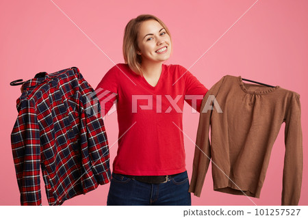 Hesitant lovely young female holds in hands two shirts, can`t choose which to buy, looks positively into camera, likes shopping, isolated over pink background. People and consumerism concept Hesitant lovely young female holds in hands two shirts, can`t choose which to buy, looks positively into camera, likes shopping, isolated over pink background. People and consumerism concept 101257527
