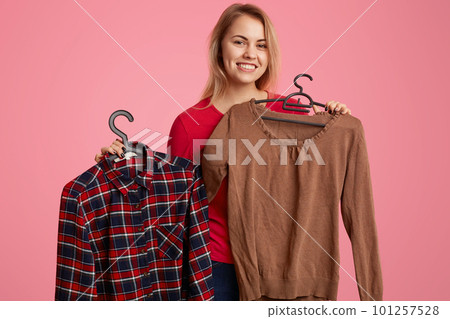 Positive optimistic woman with light hair, toothy smile, holds new clothes on hangers, rejoices successful purchases, looks directly at camera, isolated over pink background. Buying concept. Positive optimistic woman with light hair, toothy smile, holds new clothes on hangers, rejoices successful purchases, looks directly at camera, isolated over pink background. Buying concept. 101257528