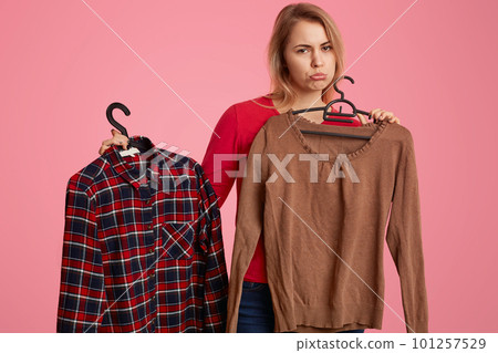 Sad dissatified Caucasian woman purses lips, holds two shirts oh hangers in shopping mall, discontent with high price, isolated over pink background. People and consumerism concept. Shopping 101257529