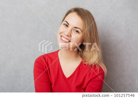 Horizontal shot of pleased pretty woman with pleasant charming smile, dressed in red sweater, expresses positive emotions, isolated over grey concrete background. Facial expressions concept. Horizontal shot of pleased pretty woman with pleasant charming smile, dressed in red sweater, expresses positive emotions, isolated over grey concrete background. Facial expressions concept. 101257546