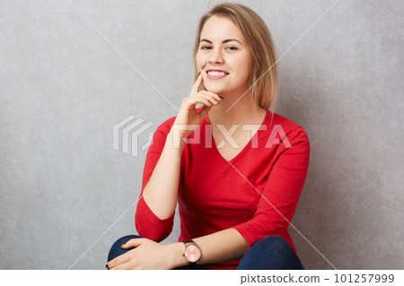 Indoor shot of pleasant looking happy female with pleasant smile on face, dressed in red sweater, poses at camera against grey background. Beautiful woman being in good mood after date with lover 101257999