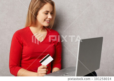Smiling female buys good in internet store, uses credit card and laptop computer, does online shopping, isolated over grey background. Cute young woman verifies her bank account on notebook 101258014