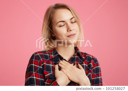 Friendly kind hearted female keeps eyes shut, hands on chest, expresses her gratitude to someone, poses against pink background. Adorable beautiful young woman relaxes indoor, being gorgeous 101258027